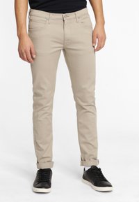 Man i beige slim-fit jeans med uppvikt fåll och svarta sneakers som står mot en vit bakgrund.