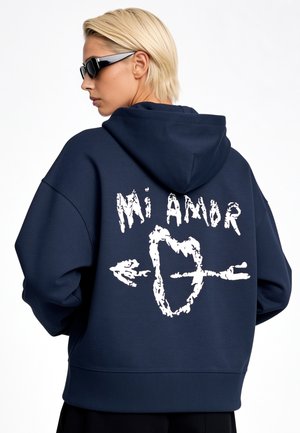 Personne aux cheveux blonds courts portant des lunettes de soleil foncées et un sweat à capuche bleu marine avec un texte blanc "Mi Amor" et un cœur traversé d'une flèche dans le dos.