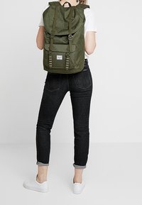 Zielony plecak z płótna z zrolowanym dnem, regulowanymi ramiączkami, przednią kieszenią i zapięciami na klamry. Noszony z czarnymi dżinsami i białymi trampkami.