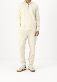 Felpa crema con zip e collo alto, abbinata a pantaloni jogger coordinati. Caratterizzata da polsini elastici e dettagli con logo ricamato.