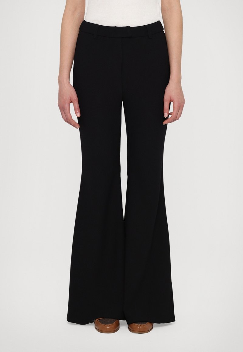 Pantalon noir taille haute à jambes larges porté avec des chaussures marron et un haut blanc, vu de face sur une personne debout.