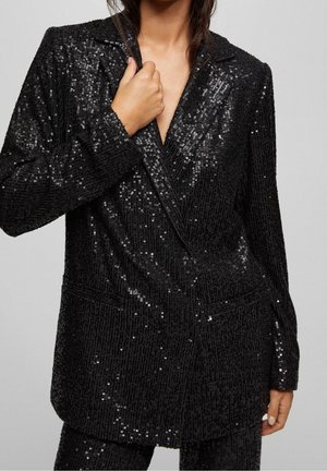 Kobieta w czarnym, cekinowym blezerze z klapami, trzymająca kołnierz jedną ręką, na jednolitym jasnym tle.