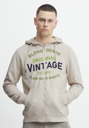 Bežna zip-up kapuljača izrađena od mješavine pamuka, s vintage printom u zelenoj i tamnoplavoj boji. Ima kapuljaču s utezima i prednje džepove.