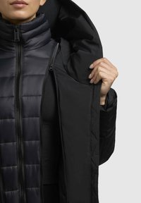 Giacca a vento nera isolata con colletto alto e cappuccio. Presenta una chiusura con zip e una texture trapuntata, che mostra la fodera interna e il tessuto liscio.