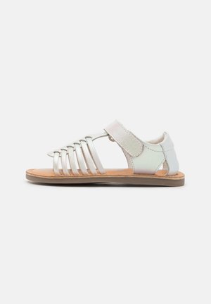 Sandalia infantil con correas metálicas blancas, diseño abierto, cierre ajustable de gancho y bucle, plantilla en tono marrón y suela texturizada.