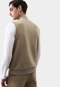 Sudadera sin mangas beige con textura suave, puños acanalados y escote redondeado, usada sobre una camisa blanca de manga larga.