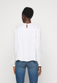 Blusa bianca a maniche lunghe con pois testurizzati, scollatura rotonda, chiusura a chiave sul retro e vestibilità morbida. Abbinata a jeans blu.