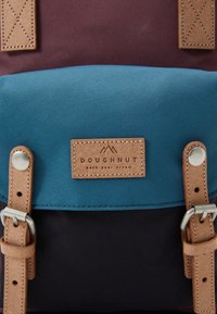 Mochila multicolorida com um topo de maroon escuro, meio teal e fundo preto. Alças de couro e um emblema de couro com a marca detalham o design.
