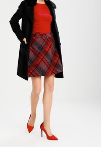 Zwarte coat met bontkraag, rode getailleerde top, rode en zwarte geruite rok en rode puntige hak schoenen. Rok heeft twee knoppen.