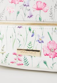 Section d'un sac blanc texturé avec une fermeture éclair dorée, imprimé floral en rose, violet et vert, et une plaque nominative rectangulaire en or.