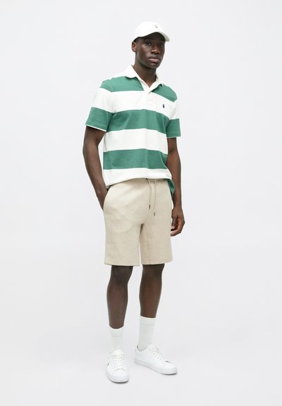 Πολυφορεμένο πράσινο και λευκό πόλο μπλουζάκι, μπεζ σορτς, λευκά αθλητικά παπούτσια και καπέλο. Χαλαρή εφαρμογή, από βαμβακερό υλικό, με διακριτό λογότυπο στο πόλο.