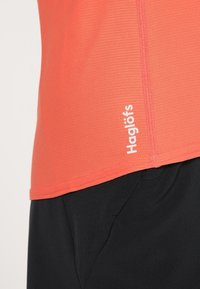Haut de sport corail avec une finition texturée, arborant un logo "Haglöfs" à l'ourlet ; associé à un tissu noir.