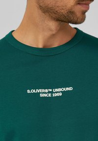 Camiseta de algodón verde con escote redondo y mangas cortas. Muestra el texto blanco "S.OLIVER® UNBOUND SINCE 1969" en la parte delantera.