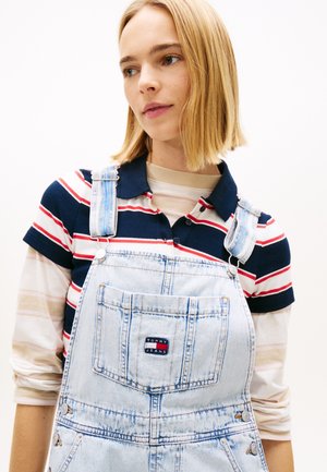 Giovane donna con capelli biondi lisci che indossa una polo a righe sotto una salopette di denim azzurro chiaro con un logo sulla tasca frontale.