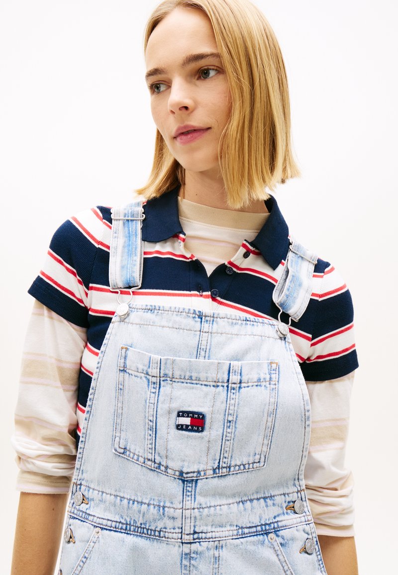 Νέα γυναίκα με ίσια ξανθά μαλλιά που φοράει ριγέ πόλο μπλούζα κάτω από ανοιχτό μπλε τζιν σαλοπέτα με λογότυπο σε μπροστινή τσέπη.