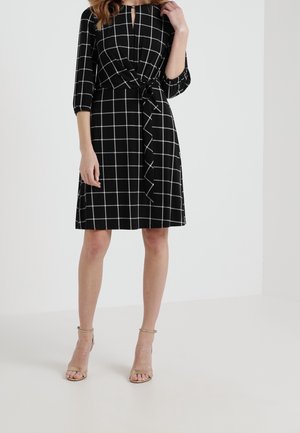 Femme portant une robe noire mi-longue à motif de grille blanche, manches trois-quarts, détail noué sur le côté, et sandales à talons hauts beiges.