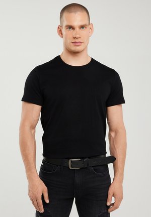 Žmogus trumpais plaukais, dėvintis prigludusią juodą marškinėlį, juodą diržą ir juodas džinsines kelnes, stovintis priešais paprastą šviesią foną.