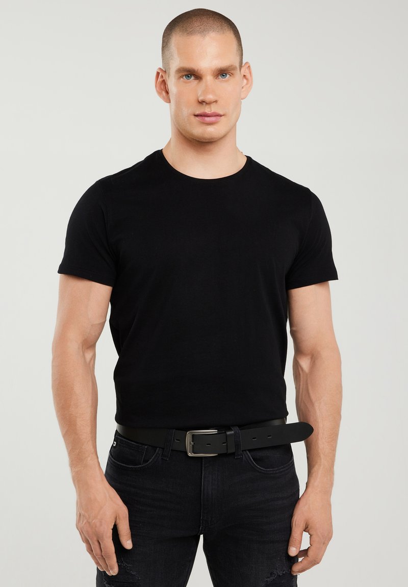 Lühikeste juustega mees, kes kannab kehale istuvat musta T-särki, musta vööd ja musti teksaseid, seisab tavalise hele tausta ees.