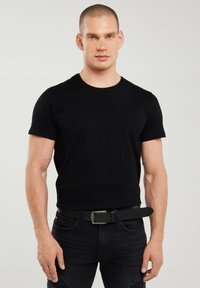 Lühikeste juustega mees, kes kannab kehale istuvat musta T-särki, musta vööd ja musti teksaseid, seisab tavalise hele tausta ees.