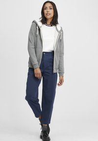 Gray zip-up hoodie with white fleece lining, worn over a white top. Blue high-waisted pants and black lace-up shoes complete the look.