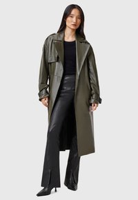 Olive green leather trench coat with wide lapels, belted cuffs, and a straight silhouette. Worn over black fitted leather pants.