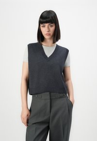 Chaleco de punto marinero con diseño de escote en V, superpuesto a una camiseta gris clara; combinado con pantalones gris de cintura alta que presentan detalles de botones.