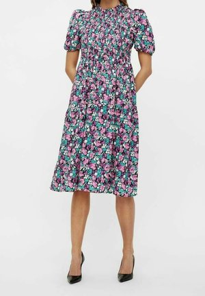 Femme portant une robe fleurie longueur genoux avec manches bouffantes, corsage smocké et talons noirs pointus sur un fond uni.