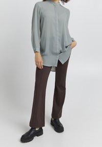 Light gray, long-sleeve blouse with a high collar and button closure paired with brown flared pants and black chunky-sole shoes.