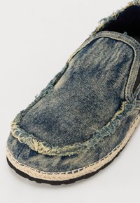 Denim slip-on shoe with frayed edges, a blue wash, jute-wrapped sole, and black rubber bottom. Features rounded toe and exposed stitching.