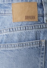 Tissu en denim bleu clair avec des détails de couture visibles ; une patch en cuir tan avec "DRYKORN" embossé. Des poches renforcées et des lignes de couture sont présentes.