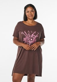 Donna che indossa un vestito a maniche corte marrone con grafica floreale rosa e la scritta "BELLEZZA" in piedi su uno sfondo bianco semplice.