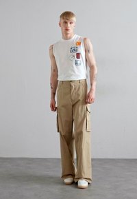 Top alb fără mâneci cu patch-uri grafice; pantaloni largi bej de tip cargo cu buzunare laterale; purtati cu adidași albi pe un podea gri.
