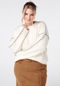 Lachende vrouw met blond haar in een hoge paardenstaart, gekleed in een witte trui met zwarte accenten en een bruine suede rok tegen een witte achtergrond.