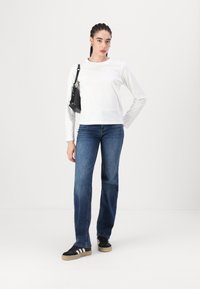Hvid langærmet skjorte, denim blå vide bukser, og sorte sneakers med hvide striber. Sort crossbody taske, neutral baggrund.
