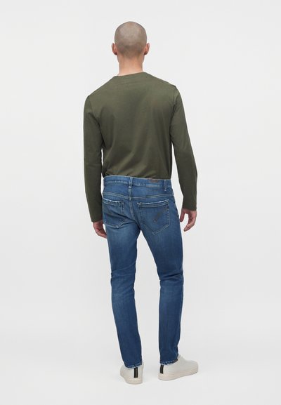 Homme portant une chemise à manches longues vert olive, un jean bleu cintré avec des poches arrière et des baskets blanches, vu de dos sur fond uni.