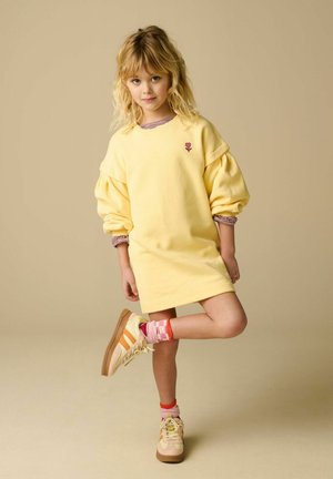 Jeune fille aux cheveux blonds, portant une robe jaune oversize, des chaussettes rayées et des baskets beige, debout avec une jambe pliée.