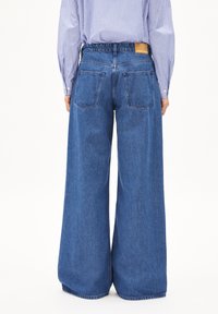 Pantaloni in denim blu a gamba larga con vita alta, fascia elastica in vita e due tasche posteriori. Tessuto leggermente texturizzato con dettagli di cucitura visibili.