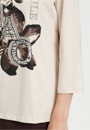 Helles beiges Baumwolloberteil mit einem floralen Druck in Dunkelbraun und Schwarz. Verziert mit silberfarbenen Nieten und einem auffälligen Grafikdesign.