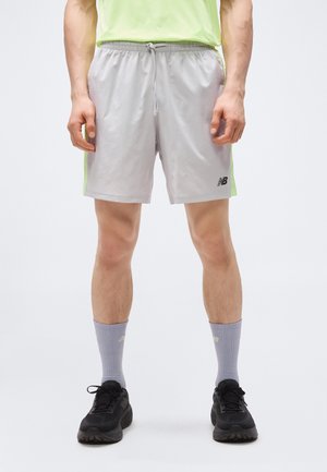 Άνδρας που φοράει ανοιχτογκρι αθλητική βερμούδα με νέον πλαϊνές λωρίδες, πουκάμισο λάιμ, γκρι κάλτσες και μαύρα παπούτσια τρεξίματος, στέκεται με τα χέρια χαλαρά.