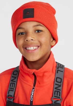 Orange ribbed beanie with a black logo patch, worn with an orange fleece jacket, featuring a high collar and gray suspenders.