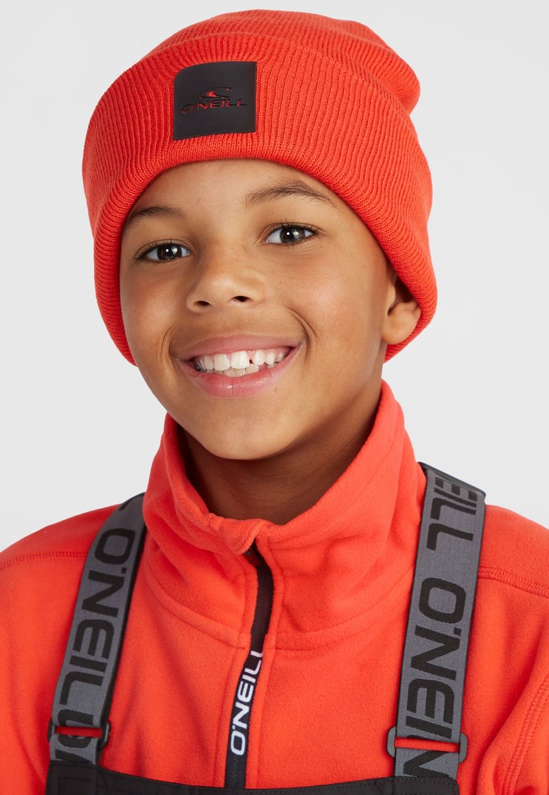 Orange ribbed beanie with a black logo patch, worn with an orange fleece jacket, featuring a high collar and gray suspenders.