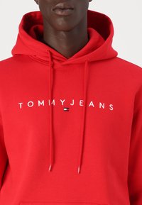 Raudona džemperis su gobtuvu, pagamintas iš medvilnės mišinio; turi kengūros kišenę, raištelį ant gobtuvo ir baltą "TOMMYJEANS" tekstą su mažu logotipu.