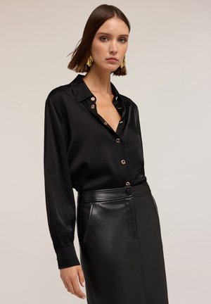 Black satin blouse with long sleeves and a collar, featuring gold buttons. Worn with a high-waisted black leather skirt.