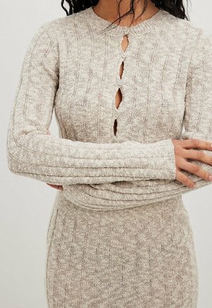 Robe beige tricotée côtelée à manches longues avec petites fermetures à boutons sur le devant et un col rond, portée les bras croisés.