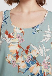 Top sin mangas floral con fondo azul claro, flores y hojas multicolores, escote redondeado y tirantes estrechos.