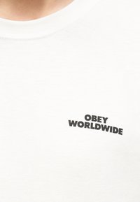 Λευκό μπλουζάκι με στρογγυλή λαιμόκοψη και μαύρο κείμενο «OBEY WORLDWIDE» τυπωμένο στο αριστερό μέρος του στήθους.