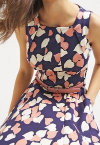 Marineblaues Kleid mit rosa und weißen herzförmigen Mustern, ausgestattet mit einem taillierten Oberteil, Falten und einem passenden Gürtel um die Taille.