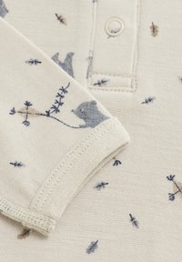 Ljus krämfärgad tyg med en mjuk textur, som har ett blått björnmönster och små blå detaljer, med en knappslå vid halsringningen.