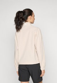 Lightweight, check-patterned jacket in beige with elastic cuffs, standing collar, and a logo detail at the back. Paired with black shorts.