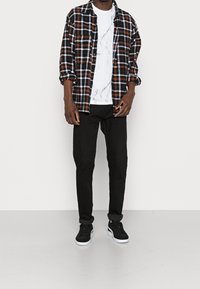 Camisa de franela en cuadros negro, blanco y naranja sobre una camiseta con patrón de mármol blanco. Jeans negros ajustados y zapatillas negras con suelas blancas.